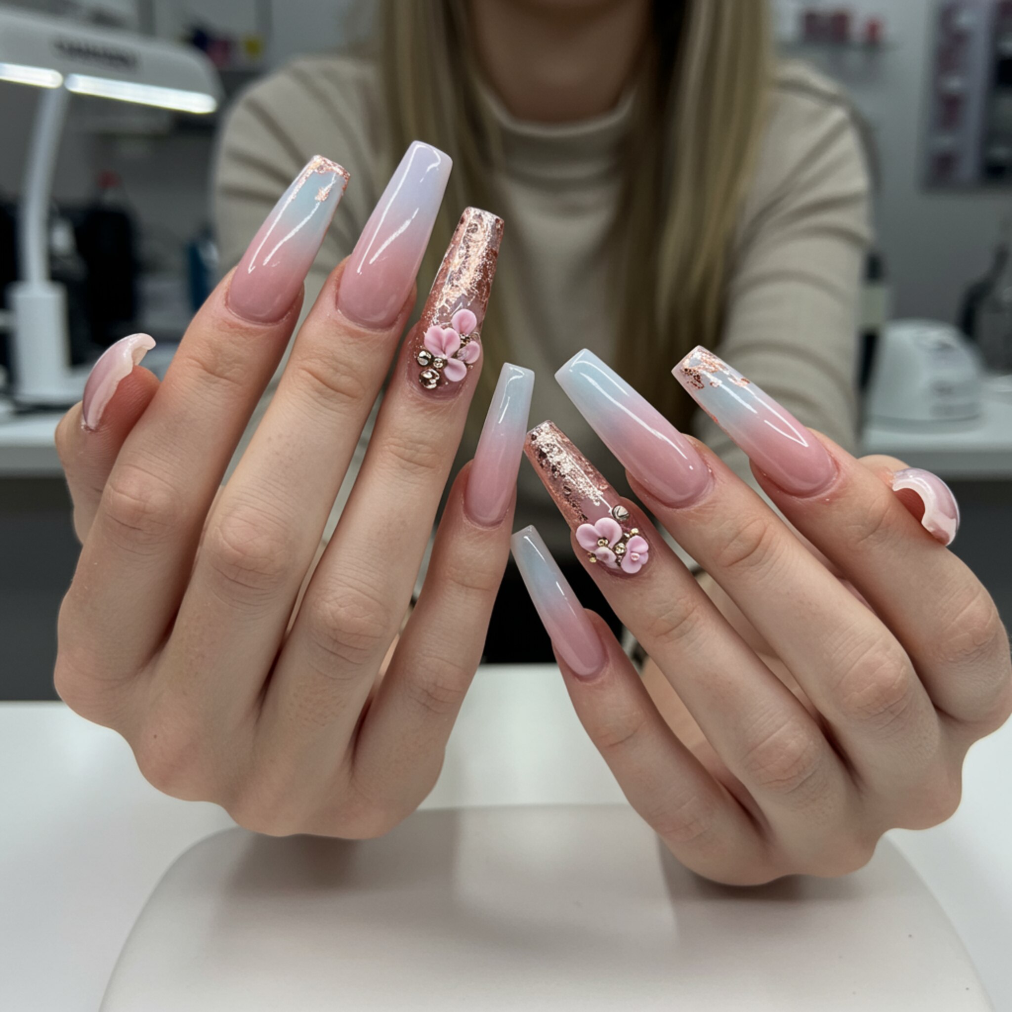 "Manos de mujer con uñas largas y esculpidas en acrílico y acrygel, diseño en tonos pastel y detalles en foil rosa dorado, destacando la calidad en un salón de manicura moderno."